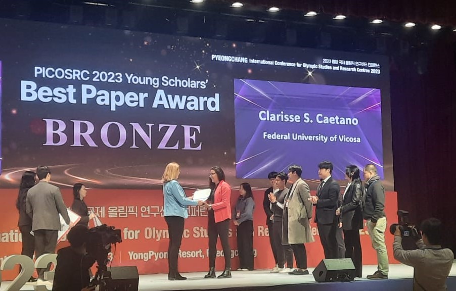 Premiação na categoria Jovens Pesquisadores  Mulher loira de azul entrega premiação a mulher morena de vermelho no palco do evento.International Conference for Olympic Studies and Research Centres, realizada em Pyeongchang, na Coreia do Sul. A pesquisa é de autoria de Clarisse Silva Caetano; da professora Doiara Silva dos Santos,  e do Augusto Fernandes Condé
